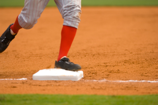 This color image is of a Baseball Player Running to First Baseball during Baseball Game. the baseball player is wearing an official baseball uniform during a baseball game. the baseball player is stepping on first base or third base after hitting the baseball. the picture includes the infield of a baseball game. the picture was taken at a live baseball game or sporting event. the lighting in natural sunlight