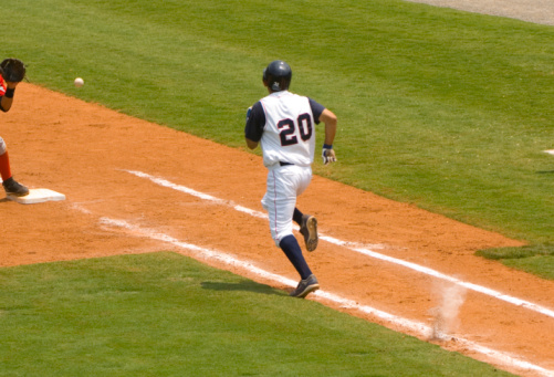 this photo is of a Baseball Player Running to First Baseball during Baseball Game. the baseball player running to first base is trying to outrun the baseball being thrown to the first baseman that is standing on first ball. the baseball player is wearing a professional baseball uniform and baseball cleats. the baseball being thrown is a professional white baseball with red stitching. the first baseman is wearing a professional baseball glove. the Baseball infield is made of lush green grass and artificial turf and dirt. the foul line is white chalk line. the and foul area is green grass or artificial turf. this photo was taken during a live baseball game or sporting event during baseball season. the lighting is natural warm sunlight during the day. 