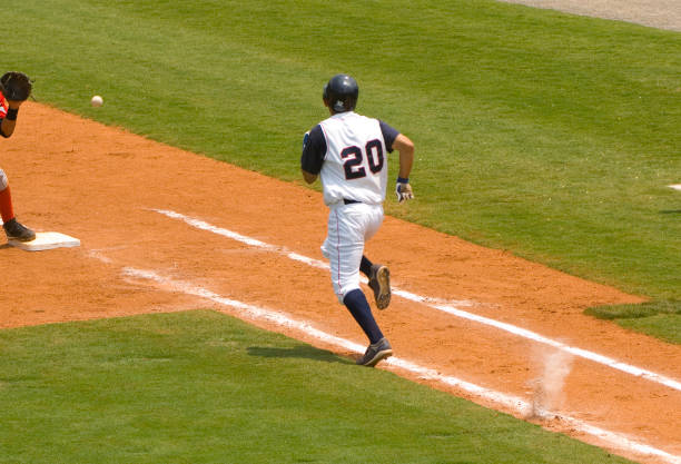 Baseball Player Running to First Baseball during Baseball Game this photo is of a Baseball Player Running to First Baseball during Baseball Game. the baseball player running to first base is trying to outrun the baseball being thrown to the first baseman that is standing on first ball. the baseball player is wearing a professional baseball uniform and baseball cleats. the baseball being thrown is a professional white baseball with red stitching. the first baseman is wearing a professional baseball glove. the Baseball infield is made of lush green grass and artificial turf and dirt. the foul line is white chalk line. the and foul area is green grass or artificial turf. this photo was taken during a live baseball game or sporting event during baseball season. the lighting is natural warm sunlight during the day.  baseline stock pictures, royalty-free photos & images