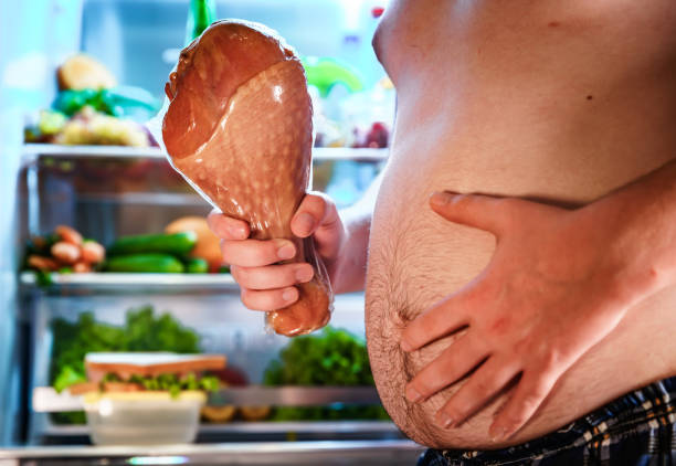 Hungry man holding a turkey leg in his hands and standing next to the open fridge. Hungry fat man holding a big turkey leg in his hands and standing next to the open fridge. Unhealthy food. thanksgiving full stock pictures, royalty-free photos & images