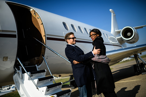 Multiethnic couple standing next to private jet airplane at the airport and hugging.