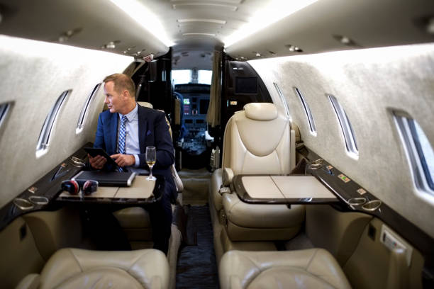 Man in private jet airplane Businessman sitting inside private jet airplane, holding digital tablet and looking outside the window. Glass of champagne, laptop computer and headphones are on the table. vehicle interior audio stock pictures, royalty-free photos & images