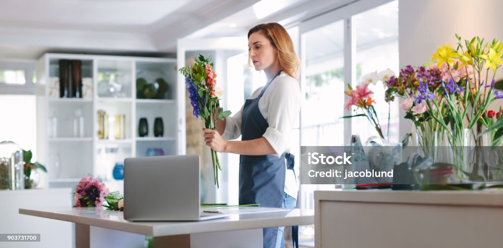 Female florist at work creating a bouquet Female florist at work creating a bouquet. Woman standing at table with laptop making a bouquet of fresh flowers. Small Business Stock Photo