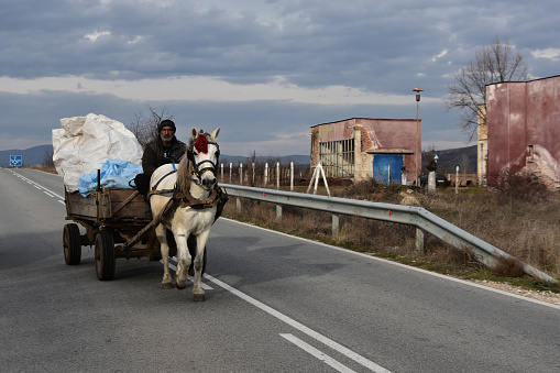 Karabunar, Bulgaria - December 16, 2017: Man from the Gypsy minority on overloaded wooden horse cart. White draft horse with red tassel. Straight asphalt road in the countryside. Ruined old buildings at background
