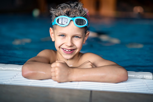 Portrait of a strong and confident little boy practicing swimming at indoors swimming pool. The boy aged 8 is smiling out of the pool into the camera. 