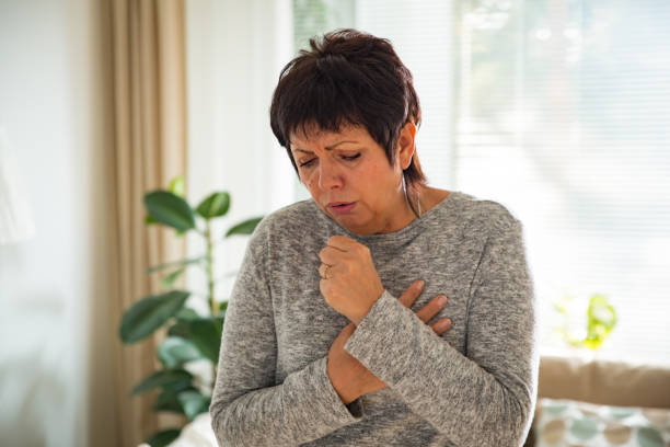 Sick mature woman with sore throat Sick mature woman with sore throat, standing in living room at home. Catching cold, having cough. coughing photos stock pictures, royalty-free photos & images