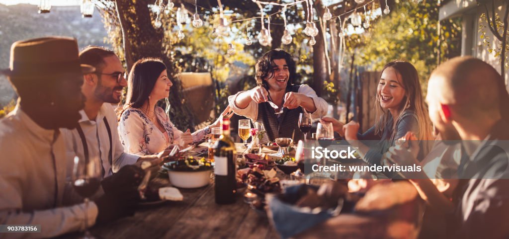 Young multi-ethnic friends dining at rustic countryside restaurant at sunset Young elegant multi-ethnic friends celebrating and having mediterranean lunch at rustic countryside mountain village house Italy Stock Photo