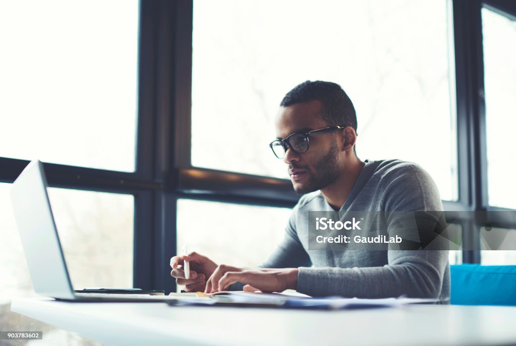Concentrated businessman checking accounting documentation in online database on modern computer connecting to wireless internet connection. Male aro american entrepreneur working on laptop indoors Business Stock Photo