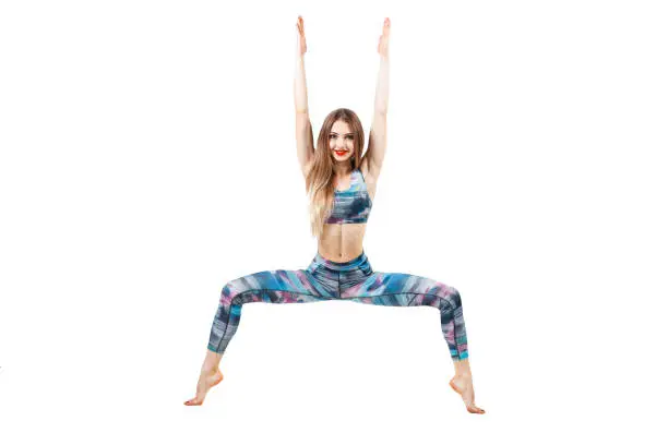 young beautiful woman in color-blue top and leggings practicing yoga, standing in Chair exercise, doing Utkatasana pose, isolated over white studio background and looking at camera