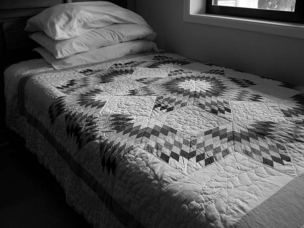 Bedroom features comfortable bed with vibrant, geometric-patterned quilt. Pale yellow walls, hardwood flooring, and sheer curtains allow natural light. Corner desk with lamp and ceiling fan complete the warm, welcoming atmosphere ideal for rest.