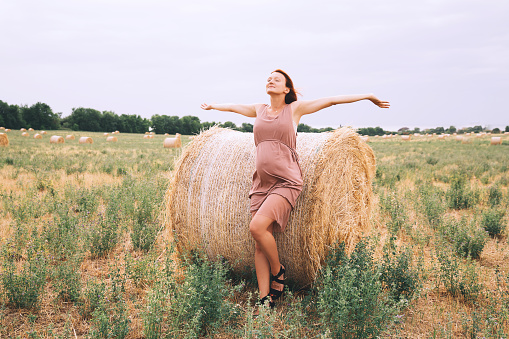Beautiful pregnant woman in dress on nature. Woman with raised arms on background of wheat field with haystacks at summer day. Photo of pregnancy, maternity, expectation. Mother waiting of a baby