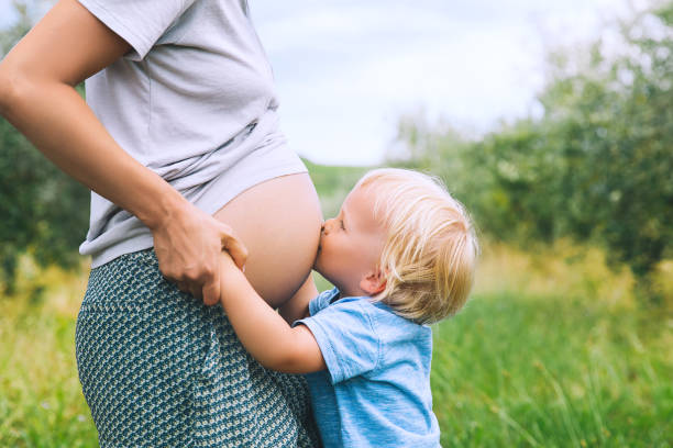 bambino ragazzo che abbraccia e bacia la pancia di sua madre incinta sullo sfondo della natura verde. - human pregnancy toddler child mother foto e immagini stock