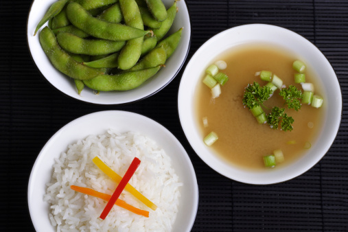 Bowls of edamame, miso soup and rice on a black reed mat