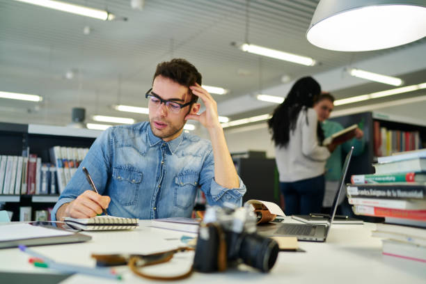 estudiante masculino se concentró en anteojos pensando en nueva tesis para escribir cursos haciendo notas en bloc de notas sentado en la biblioteca de la universidad con diferentes libros en la búsqueda de literatura útil - old cars audio fotografías e imágenes de stock