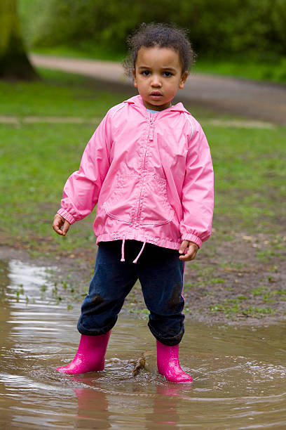A little mixed race girl in pink coat and boots standing in a muddy puddle