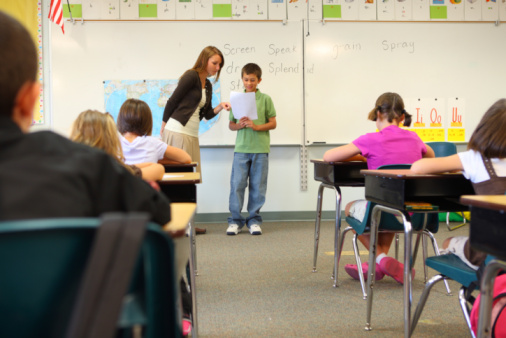 A pregnant African-American teacher standing in the front of a classroom at the whiteboard with two of her elementary students. An 11 year old boy is standing next to her, reading aloud from his notebook. A multiracial girl is sitting at a table watching and listening.