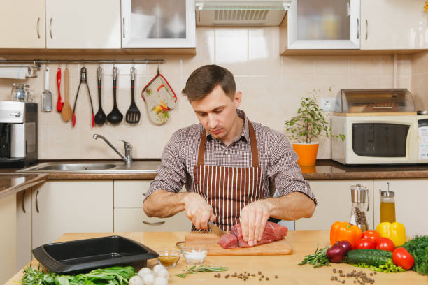 istock Handsome caucasian young man in apron sitting at table with vegetables, cooking at home preparing meat stake from pork, beef or lamb, in light kitchen with wooden surface, full of fancy kitchenware. 904068964
