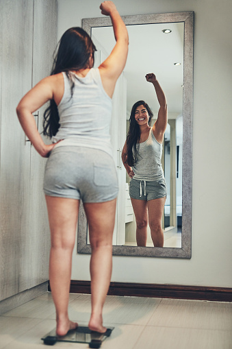 Shot of a young woman cheering while weighing herself on a scale at home