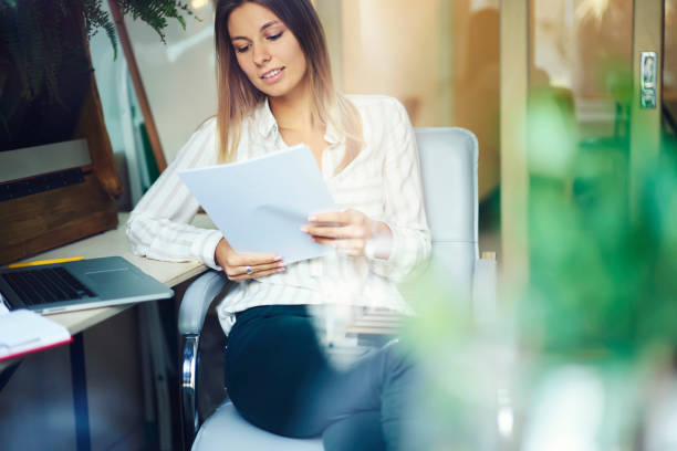 istock Intelligent female student in casual wear reading coursework written on papers with mock up area for your advertise sitting on chair at desktop with laptop computer device in office interior 904127654