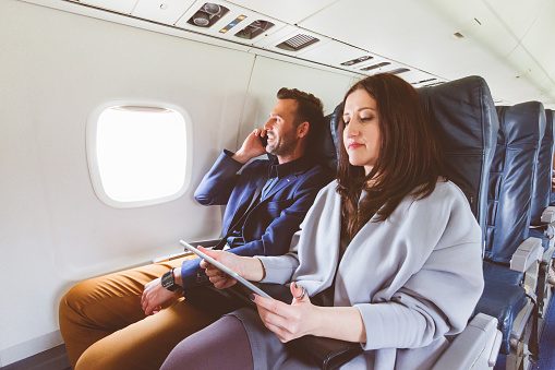 Interior of airplane with couple sitting and looking outside the window. People traveling by plane with digital tablet and mobile phone in hand.