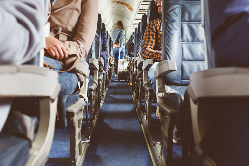 Interior of airplane with people sitting on seats. Passengers with suitcase in aisle looking for seat during flight.
