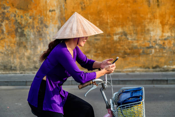 Vietnamese woman using mobile phone on a bicycle, old town in Hoi An city, Vietnam Vietnamese woman using mobile phone on a bicycle in old town in Hoi An city, Vietnam. Hoi An is situated on the east coast of Vietnam. Its old town is a UNESCO World Heritage Site because of its historical buildings. vietnamese culture photos stock pictures, royalty-free photos & images