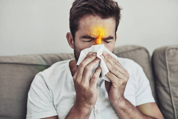No I can't get sick today Shot of a uncomfortable young man holding a tissue over his nose and sneezing while being seated on a couch cold virus stock pictures, royalty-free photos & images