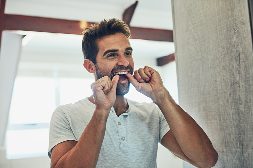 Shot of a cheerful young man flossing his teeth while looking at his reflection in the mirror at home