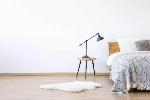 Empty white bedroom with wooden floor and bed with blanket, stool with a lamp and fur rug