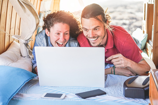 Happy couple watching videos on laptop computer inside minivan at sunset - Young people having fun on alternati summer vacation - Travel, technology and van lifestyle concept - Focus on man face