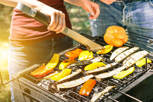 młoda kobieta gotująca warzywa na wegetariański obiad z grilla na świeżym powietrzu - para grillowania papryki i bakłażanów do grillowania - koncepcja wegańskiego i zdrowego stylu życia - miękkie skupienie się na dolnych szczypce do grillowania - couple dinner summer sunlight zdjęcia i obrazy z banku zdjęć