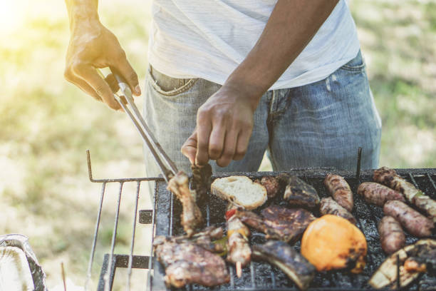 afro amerikaner kochen fleisch am grill - chef grill im park im freien - konzept des essens im freien während der sommerzeit - vintage retro-filter mit sonne halo flare einige würstchen aufsetzen - barbecue chicken stock-fotos und bilder