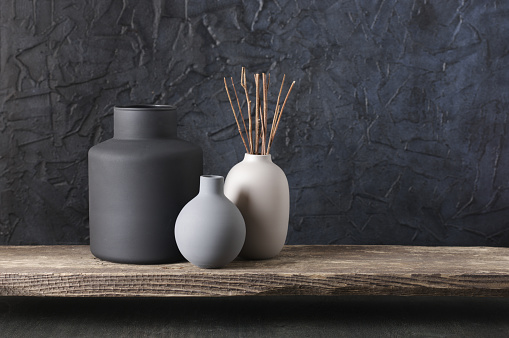 Neutral colored vases with wood sticks on distressed wooden shelf against rough plaster black wall. Home decor.