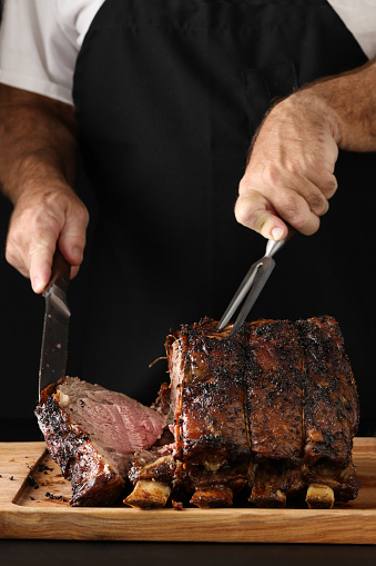 A close up vertical photograph of a chef slicing off the end piece of a freshly cooked prime rib. Isolated in black.