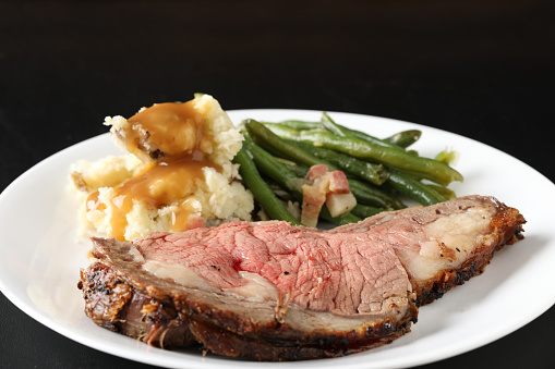 A high angle close up horizontal photograph of an   oblong plate with a medium rare cut of prime rib, mashed potatoes with gravy and steamed green beans. Isolated in black