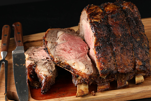 A close up vertical photograph of a freshly roasted and partially sliced prime rib plus the  carving utensils on a wooden carving board.Isolated in black.