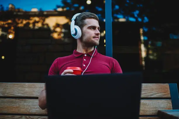 Photo of Cheerful young man 20s with modern headphones on head looking away sitting on bench with laptop device.Positive meloman with coffee in hand and computer listening electronic music via earphones