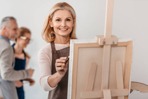 beautiful mature woman smiling at camera while painting on easel at art class beautiful mature woman smiling at camera while painting on easel at art class art class photos stock pictures, royalty-free photos & images