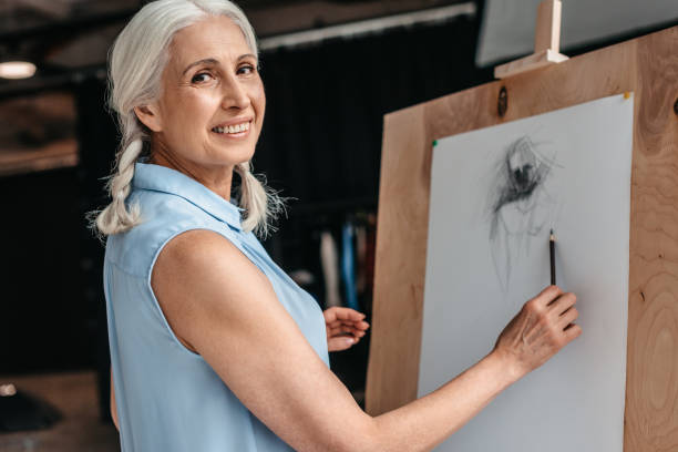 beautiful senior woman smiling at camera while drawing with pencil on easel at art class