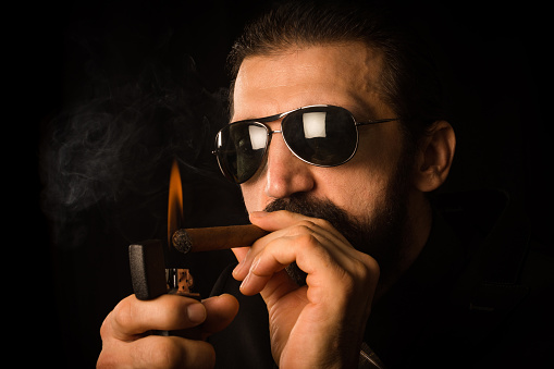 The elegant gentleman in sunglasses is igniting his cigar with zippo on black background.