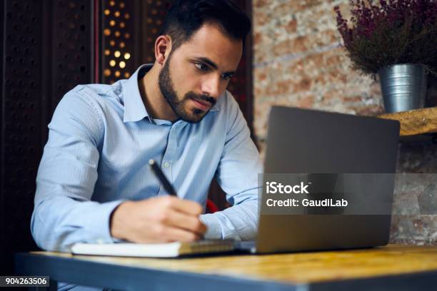 Pensive Handsome Bearded Male Owner Of Trading Company Searching Information About Marketing Agency Noting Their Contacts To Arrange Formal Meeting Sitting In Coworking Space With Laptop Computer Stock Photo - Download Image Now