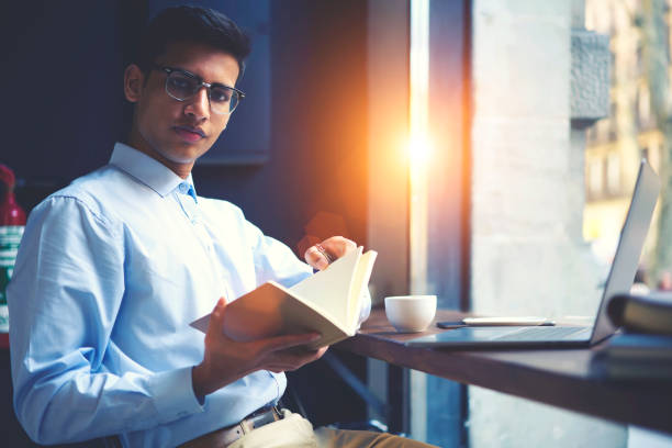 istock Talented handsome young male hipster guy sitting in cafe indoors with modern digital devices to prepare homework task on literature lesson while reading favorite detective story analyzing text 904265664