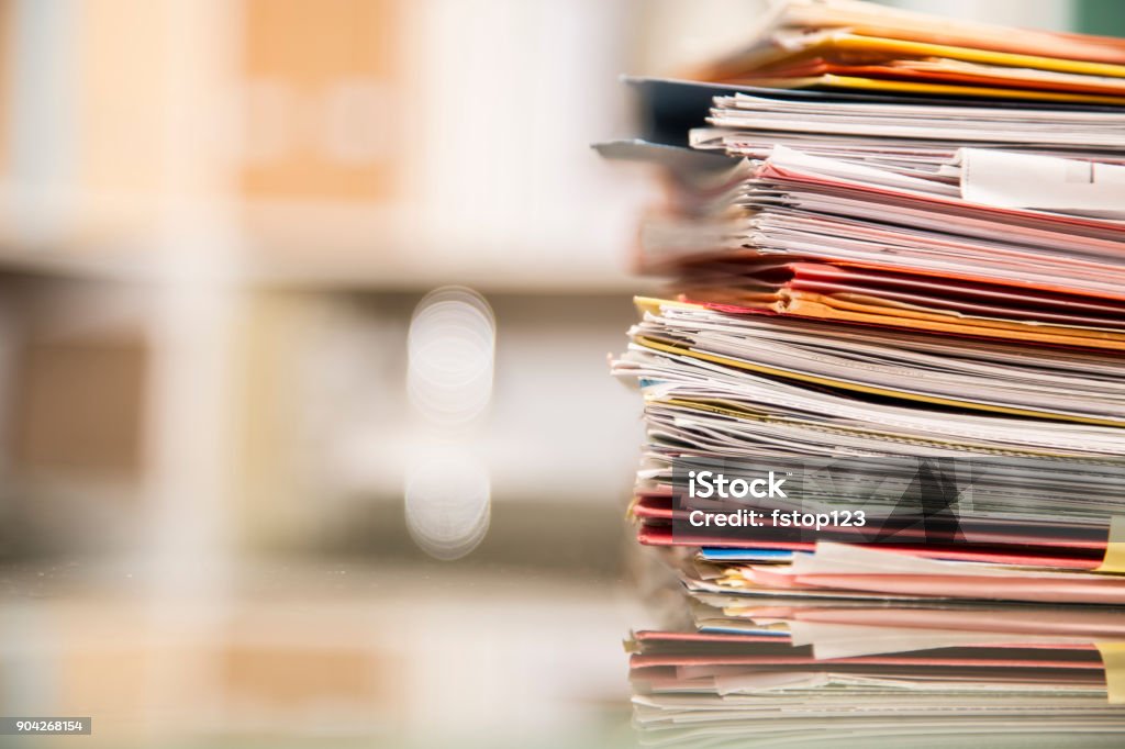 Large stack of files, documents, paperwork on desk. Large stack of file folders, documents, paperwork piled on glass top desk in office.  Bookshelves in background. Document Stock Photo