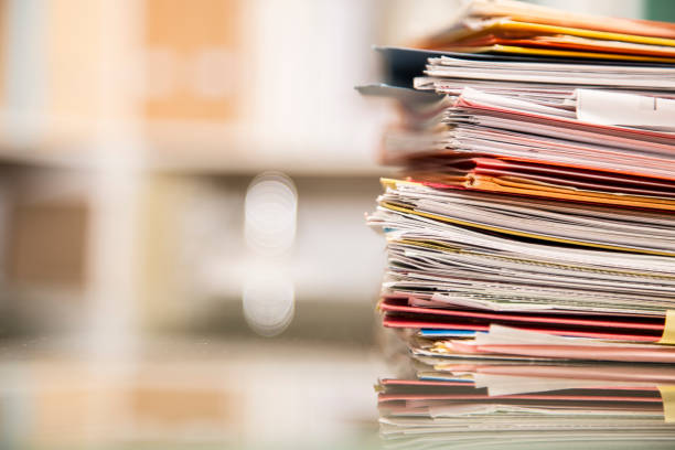 Large stack of files, documents, paperwork on desk. Large stack of file folders, documents, paperwork piled on glass top desk in office.  Bookshelves in background. bureaucracy stock pictures, royalty-free photos & images