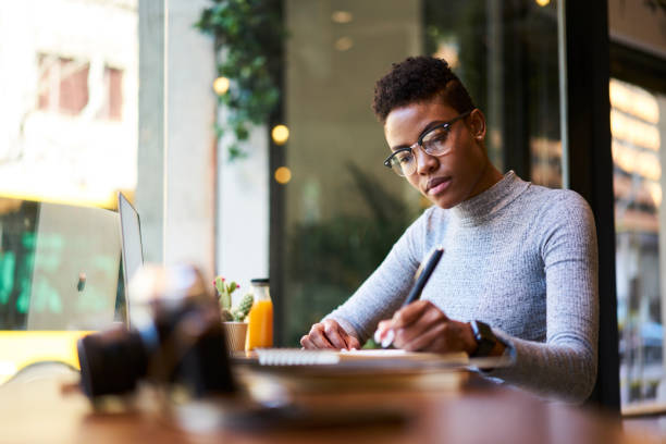Concentrated afro american experienced woman journalist in glasses creating article working in coffee shop, skilled young female graphic designer making sketch in notebook for new startup project