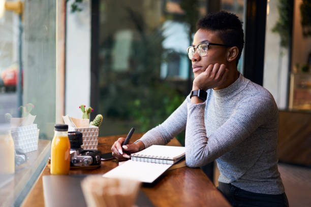 Concentrated young afro american girl student working on coursework project writing down ideas into notebook, professional female manager in eyewear making accounting report sitting in cafe indoors