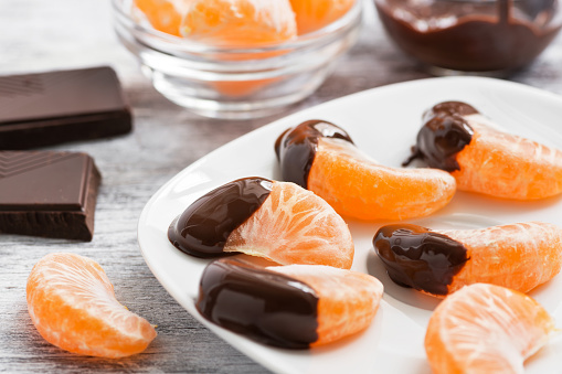 Slices of tangerine in chocolate served on a white plate, white wooden textured background.Selective focus