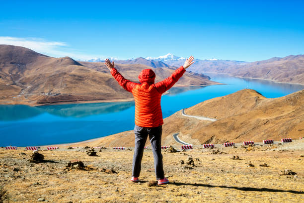 Rear view of female hiker with arms raised standing at a height of 4900 m in Tibet. In the background is seen Yamdrok Lake (Yamdrok Yutso in Tibetan dialect) and the Noijinkangsang peak (7206 m) in Himalaya. Yamdrok Lake is situated about 100 km from Lhasa, the capital of Tibet.