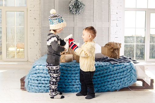 Children brother and sister standing and playing at home in the bedroom near bed with boxes, gifts on the background of Christmas decor a sunny day.Wear a warm knitted woolen garment cap and mittens.