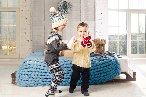 Children brother and sister standing and playing at home in the bedroom near bed with boxes, gifts on the background of Christmas decor a sunny day.Wear a warm knitted woolen garment cap and mittens.